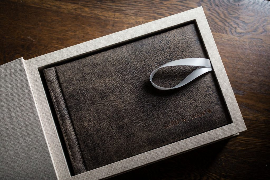 A dark brown leather-bound book with a textured cover sits inside a beige fabric box on a wooden surface, with a white satin ribbon looped on top of the book for easy removal.