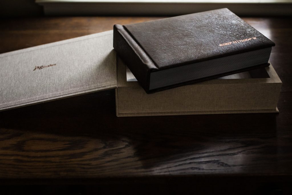 A dark brown leather-bound book rests on top of an open beige fabric-covered box, both placed on a wooden table under soft natural light.