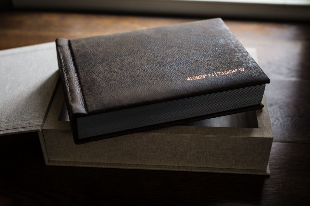 A closed, dark brown leather-bound book with gold embossed text rests on top of a light-colored, textured box on a wooden surface, with light coming from a window in the background.