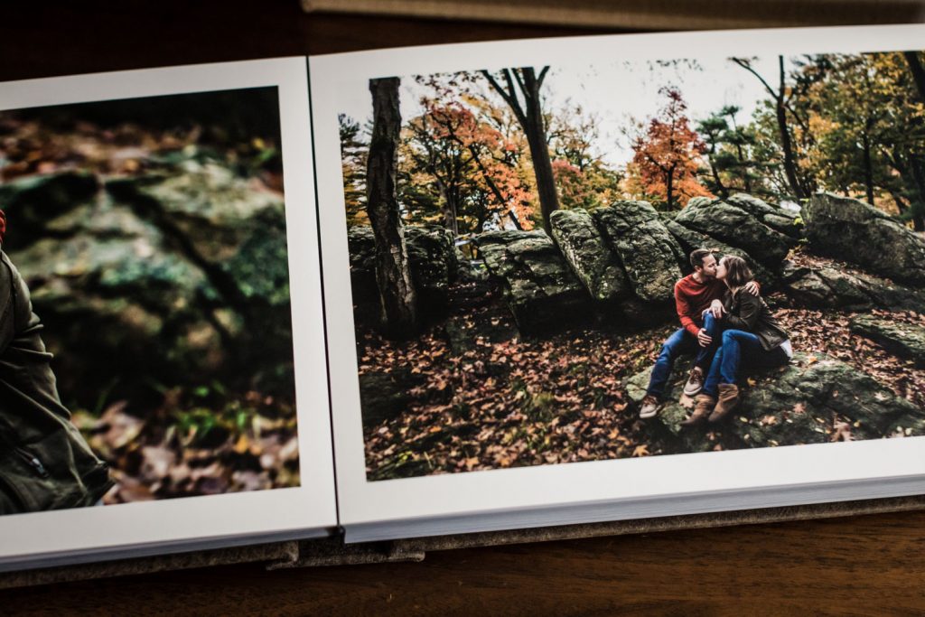 An open photo album showing a couple sitting on large rocks in a forest during autumn, surrounded by trees with fall foliage, sharing an intimate moment.