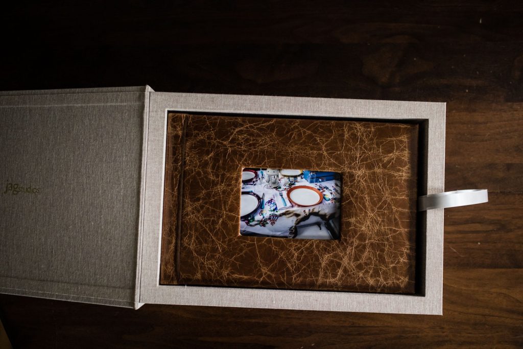 A beige photo box with a ribbon is open, revealing a single photograph of a decorated dining table set with plates, glasses, and festive decorations, resting on textured brown material inside the box.