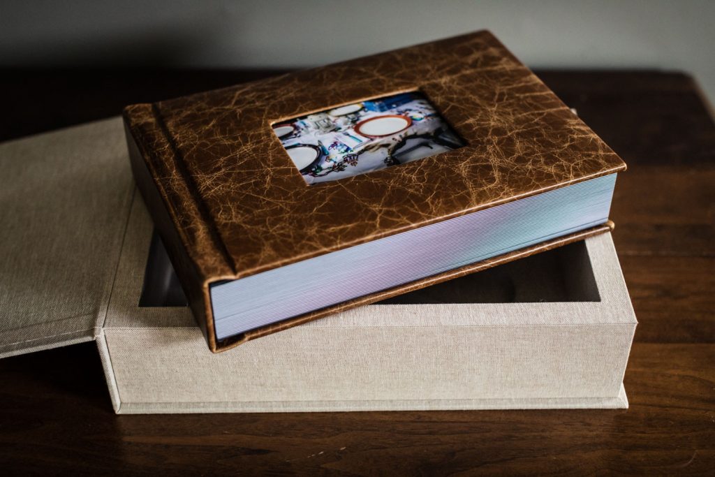A brown leather-bound photo album with a picture inset on the cover rests partially inside an open beige fabric-covered box on a wooden surface.