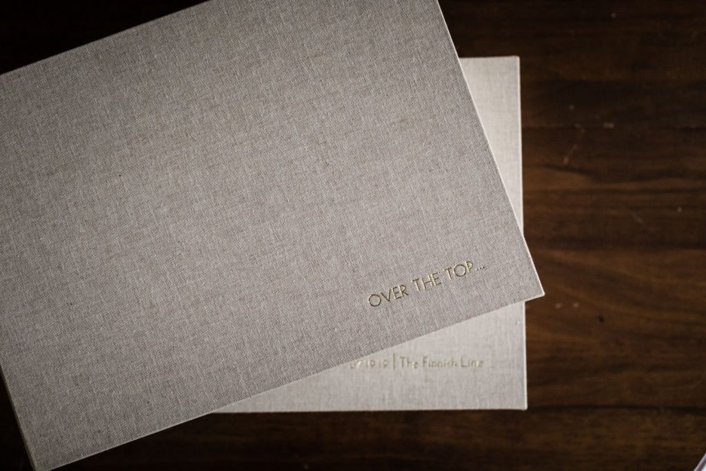 A close-up of a beige textured box with gold-embossed text reading OVER THE TOP on the lid, placed on top of another similar box, both on a dark wooden surface.