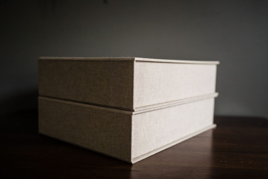 Two beige fabric-covered photo album boxes stacked on a dark wooden table against a plain, dark background.