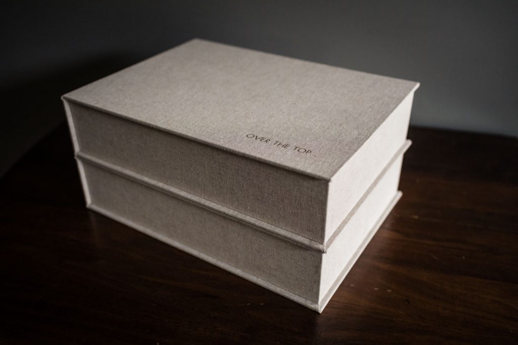 A beige rectangular box with the words OVER THE TOP printed on the lid sits on a dark wooden surface against a plain background.