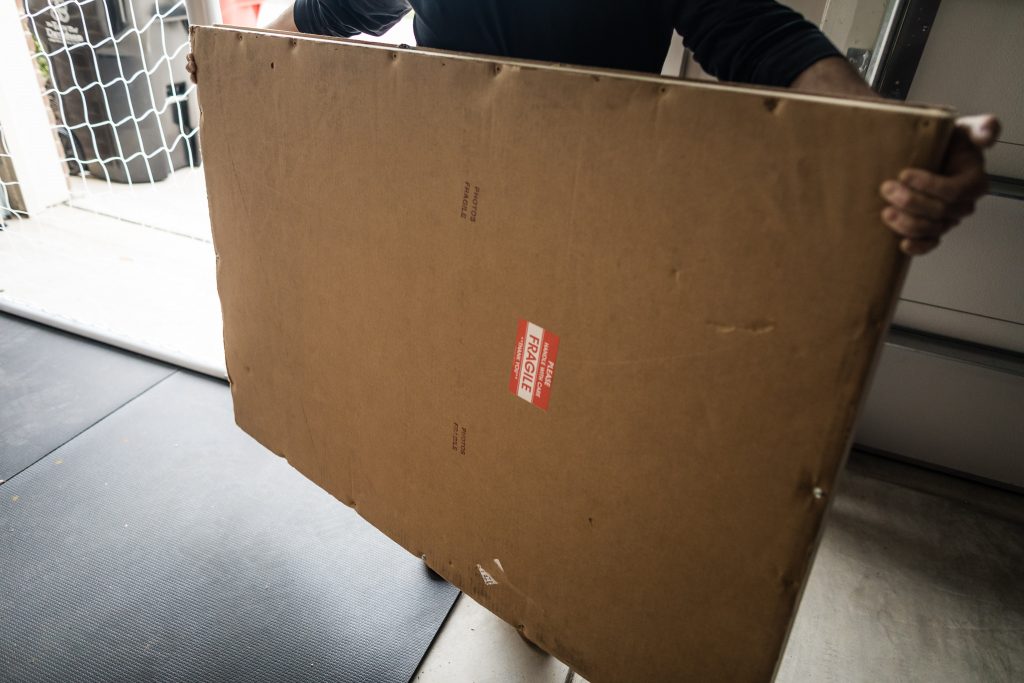 A person holds a large, rectangular cardboard box labeled FRAGILE in red text, standing in a garage near a mat and a netted door.