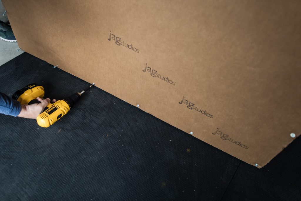 A person uses a yellow power drill to secure screws into a brown board labeled jag studios on a black textured floor.
