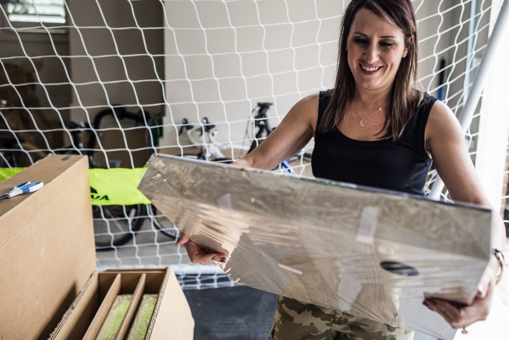 A woman smiles while holding a large, wrapped rectangular package. She stands near open boxes in a room with a net and bicycles in the background.