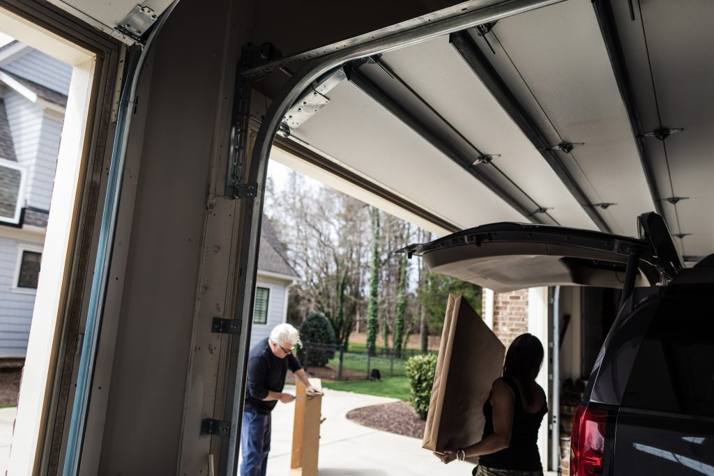 A person stands outside assembling a box while another person carries a large cardboard box toward an SUV parked in a garage with the trunk open. Trees and houses are visible in the background.