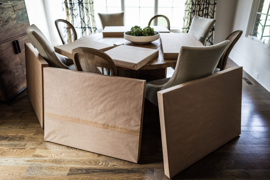 A dining room with a wooden table surrounded by chairs; framed pictures wrapped in brown paper rest on and around the table, suggesting packing or moving. Sunlight comes through large windows with curtains.