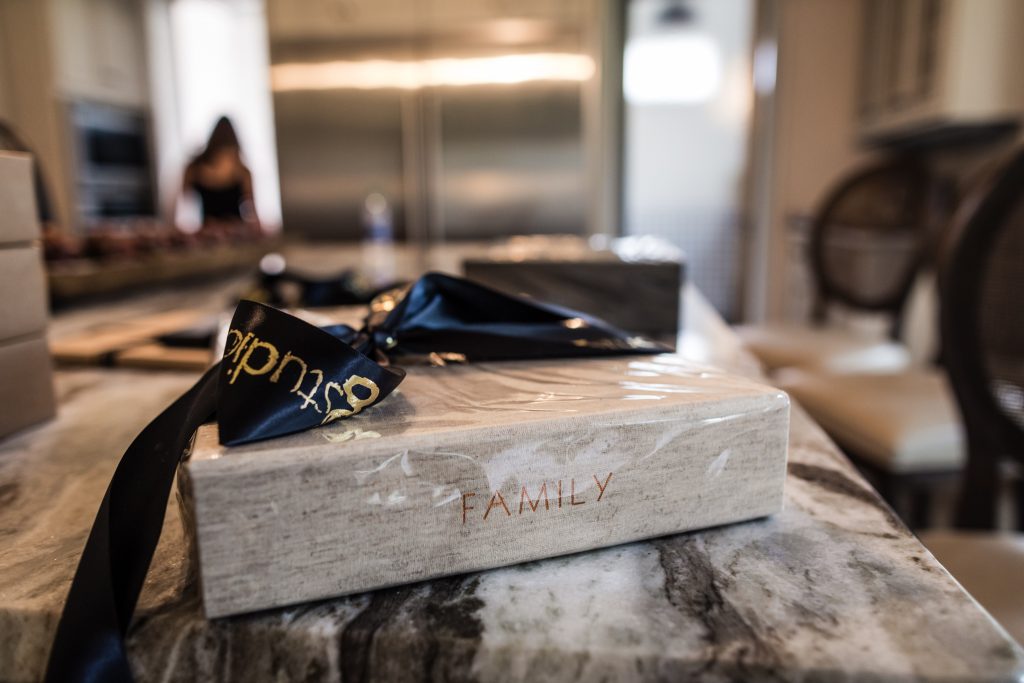 A beige box labeled FAMILY sits on a marble countertop, wrapped in plastic with a black and gold ribbon. Blurred kitchen and a person in the background.