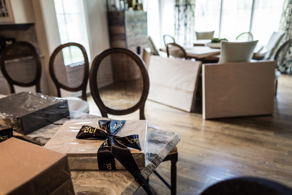 Several wrapped gifts, one with a black ribbon, sit on a marble table in a bright, modern dining room with wooden floors and large windows.