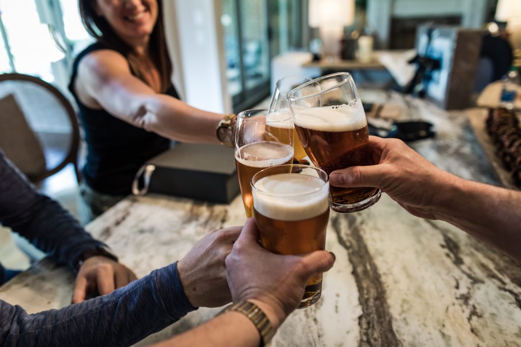 Four people clink glasses of beer together over a marble countertop, celebrating in a casual indoor setting. One woman smiles in the background, arm extended to join the toast.