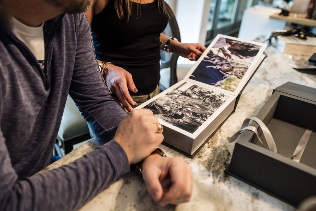 Two people looking through a photo album on a kitchen counter, with one person pointing at pictures and a gift box with a ribbon nearby.
