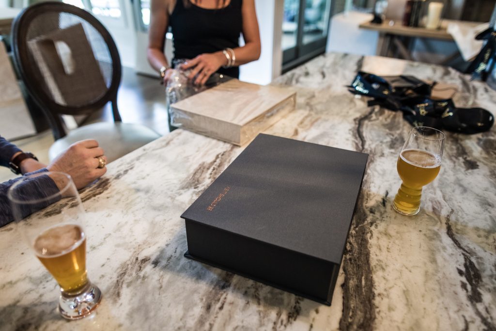 Three people sit around a marble table with two glasses of beer, a closed black box, and a large book. One person is opening a plastic bag; only their arms and hands are visible.