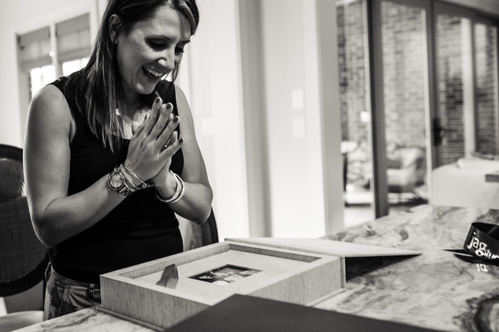 A woman stands at a table, smiling with delight and clasping her hands together as she looks at an open box containing photos. The scene appears indoors, with soft light filling the room.