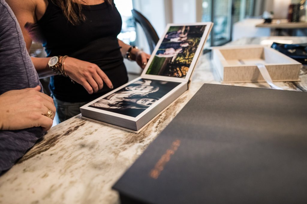 Two people stand at a marble countertop, looking at an open photo album filled with black and white and color photos. A box and a closed album are also on the counter. Only their hands and torsos are visible.