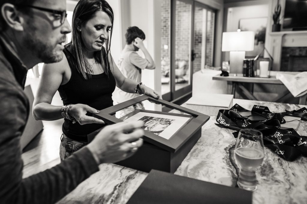 A woman and a man examine a framed photo on a kitchen counter. A boy stands in the background. The setting is a modern, well-lit home with various objects, including a glass of beer, on the marble countertop.