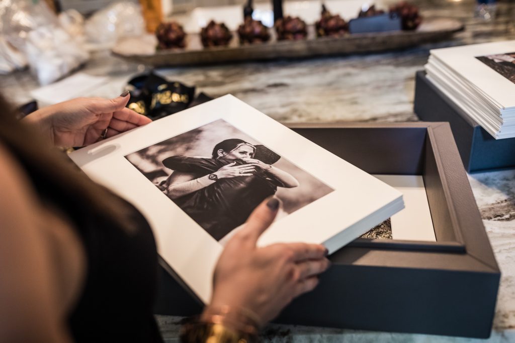A person holds a box of photo prints, with the top print showing two people embracing. The box rests on a marble countertop, and other photos and objects are visible in the background.