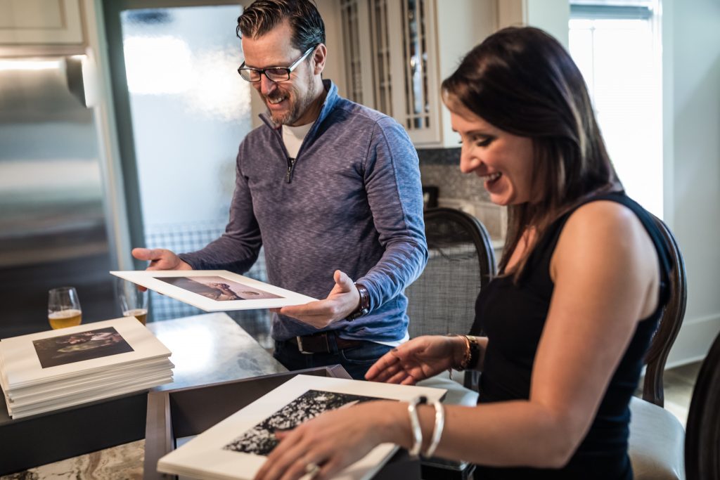 A man and a woman smile and look at printed photos together in a kitchen, standing at a table covered with photo prints and a drink in the background.