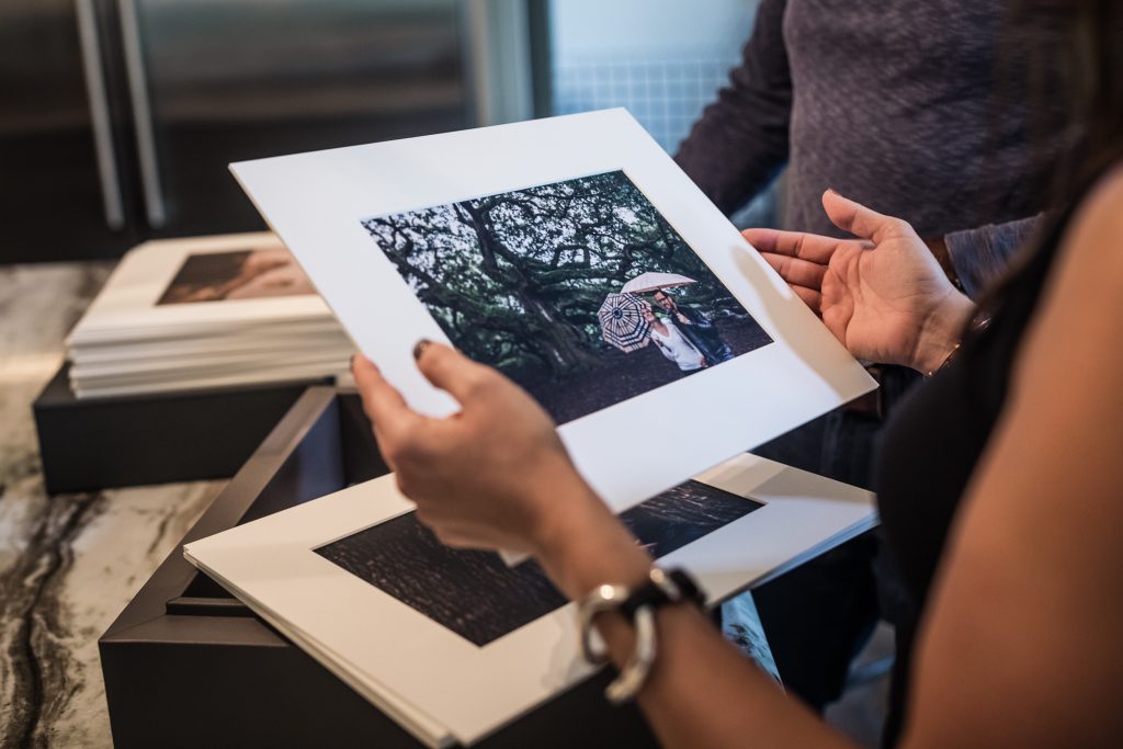 A person holds a photo print of someone with an umbrella among trees, while another person stands nearby. More photo prints and a box are on the marble countertop.