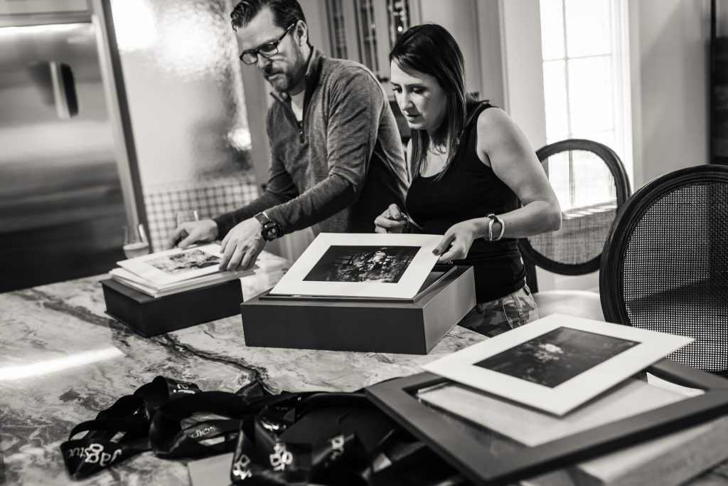 A man and a woman stand at a kitchen island looking at framed photographs. Several photos and picture frames are spread out on the counter in front of them. The scene is in black and white.