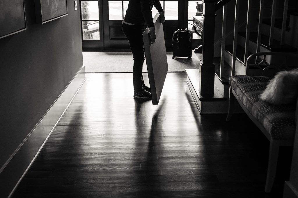 A person stands in a hallway holding a large, flat object, possibly a painting or board, with sunlight streaming through glass doors in the background. The scene is in black and white.
