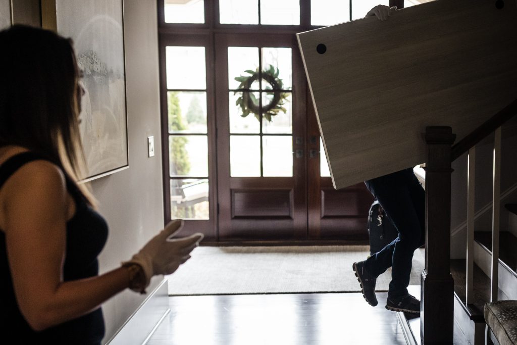 A person wearing gloves stands in a hallway while two movers carry a large piece of furniture up the stairs near a front door with a wreath.