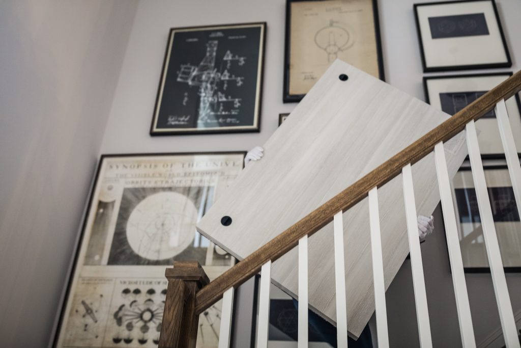 A person carries a large rectangular tabletop up a staircase, obscured by the object. The stairway wall is decorated with framed scientific and technical drawings.
