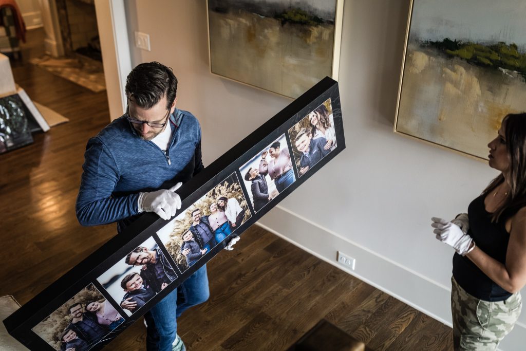 A man and a woman wearing white gloves handle a long framed collage of family photos in a living room with wooden floors and abstract art on the walls.