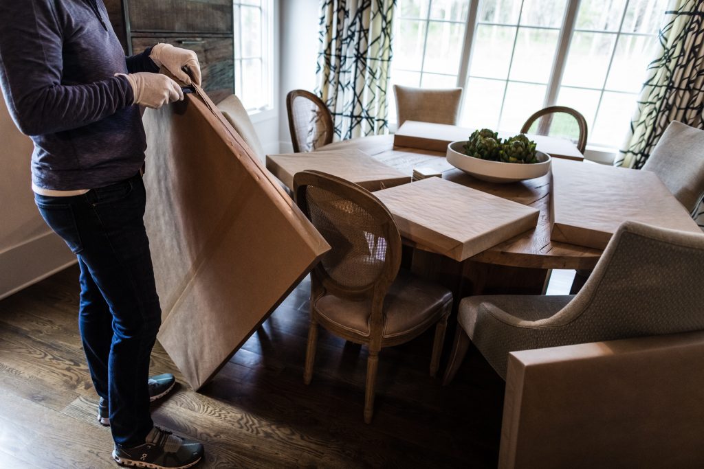 A person wearing gloves and casual clothes covers a dining table and chairs with protective brown paper in a bright room with large windows and patterned curtains.