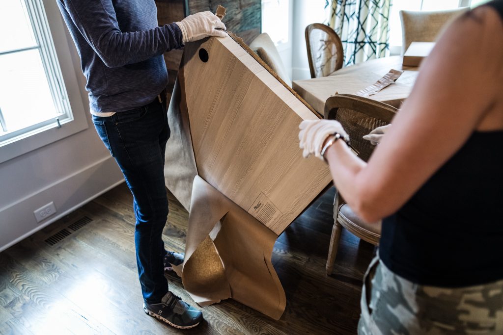 Two people wearing gloves unpack a large, flat cardboard box in a dining room with wooden floors and a table with chairs nearby. Natural light enters through a window on the left.