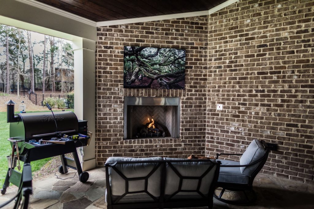 A cozy outdoor patio with brick walls features a lit fireplace, a painting of a large tree above it, a sofa and chair, and a grill beside an open doorway leading to a grassy yard with trees in the background.