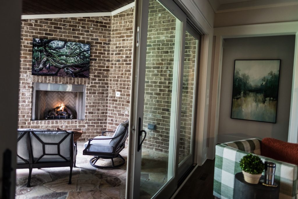 A cozy patio with a brick fireplace, TV mounted above it, and cushioned chairs is viewed through a glass sliding door; modern art and a plant decorate the adjacent indoor space.
