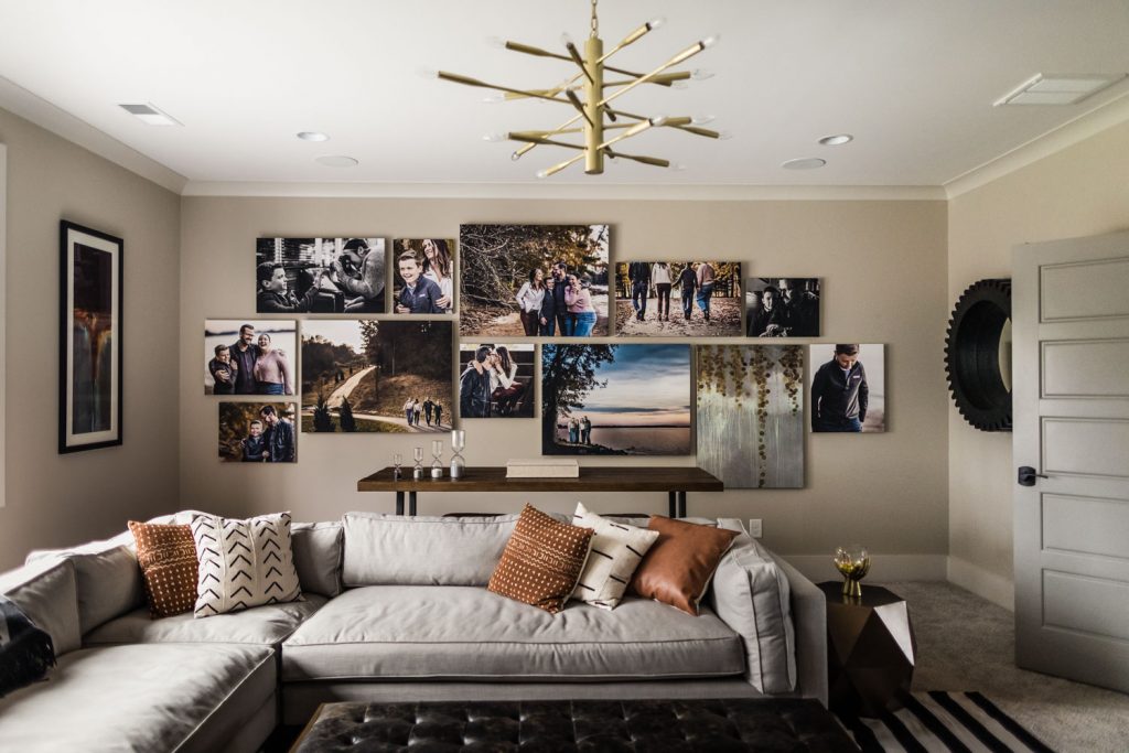 A modern living room with a gray sectional sofa, patterned pillows, a black ottoman, and a gallery wall of family photos above a console table, beneath a gold chandelier.