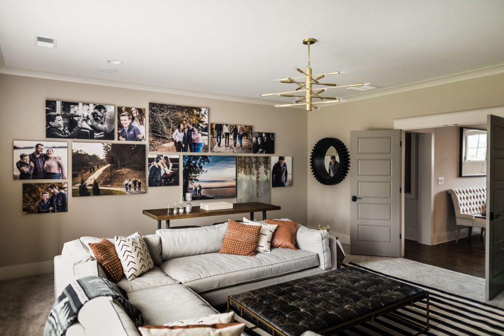 A modern living room with beige walls, a large sectional sofa with patterned pillows, a black coffee table, and a gallery wall displaying family photos. A round mirror and gold chandelier add contemporary touches.