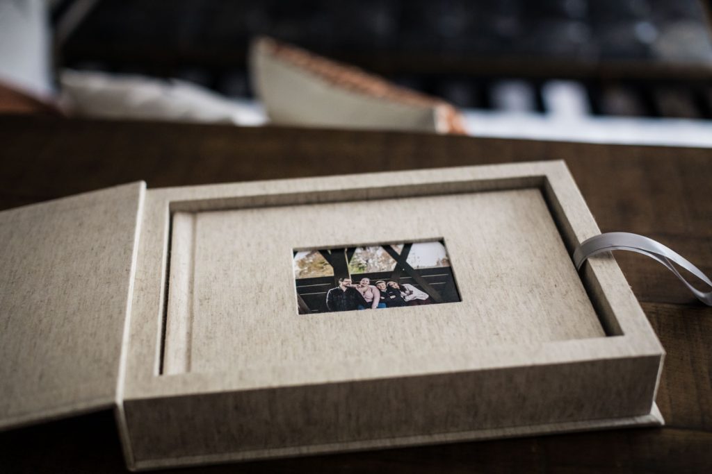 A beige photo box lies open on a wooden surface, revealing a small photo of a group of people inside the lid, with a white ribbon attached to the box for easy opening.