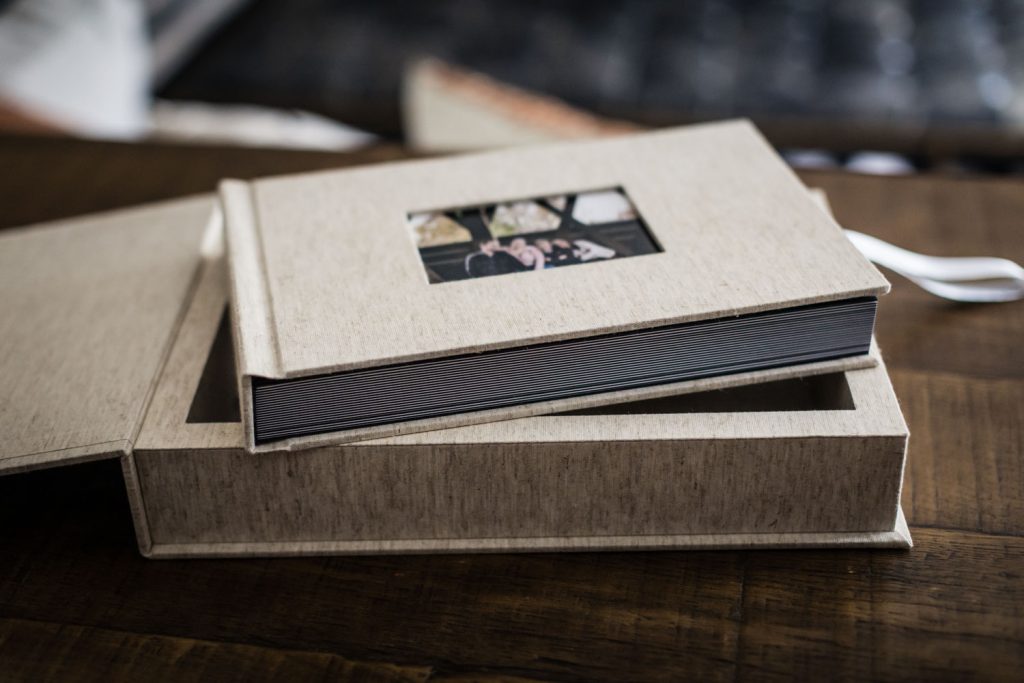 A beige photo album with a small cover photo rests partially inside an open matching box on a wooden table. The albums pages are visible from the side, and a white ribbon is attached to the box.