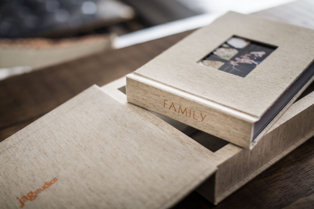 A beige photo album with FAMILY embossed on the cover sits partially inside a matching box on a wooden surface. The album cover has a small window displaying a family photo.