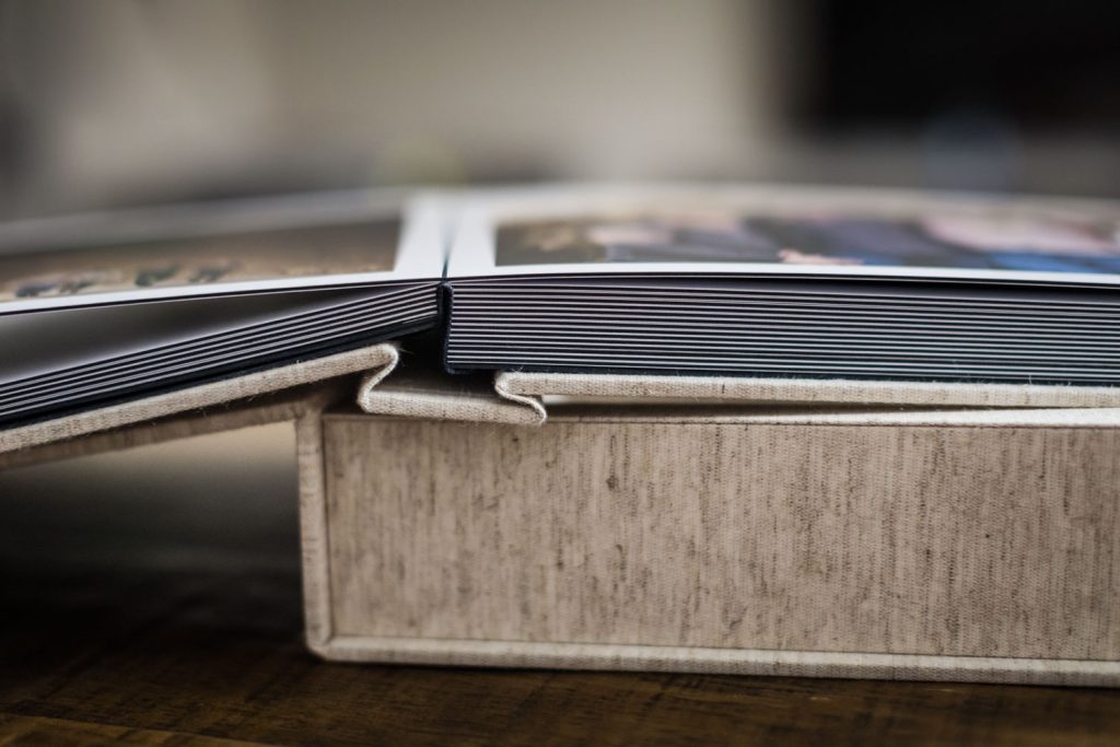 Close-up of an open photo album with thick, glossy pages lying on a textured, beige cover, resting on a wooden surface. The album is partially open, showing the binding and page edges.