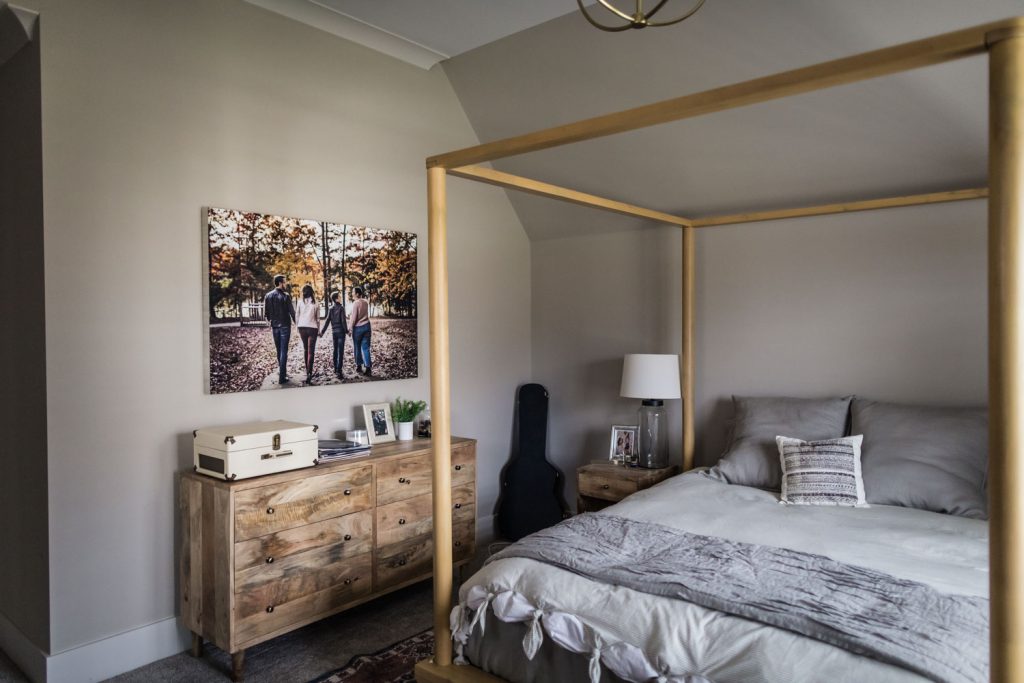 A cozy bedroom with a wooden four-poster bed, gray bedding, and a matching wooden dresser. A family photo, lamp, white record player, and guitar decorate the space. The walls are painted light gray.