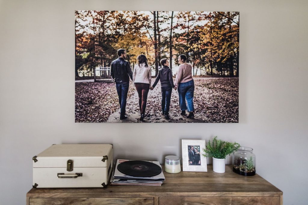 A wooden console holds a record player, vinyl, candle, framed photo, and small plants. Above, a large photo shows four people walking hand-in-hand on a tree-lined path with autumn leaves.
