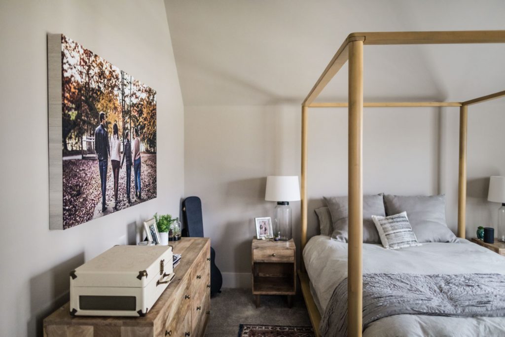 A cozy bedroom with a four-poster bed, gray bedding, wooden nightstands, a dresser with decor, a guitar by the wall, and a large photo of three people walking outdoors hanging above the dresser.