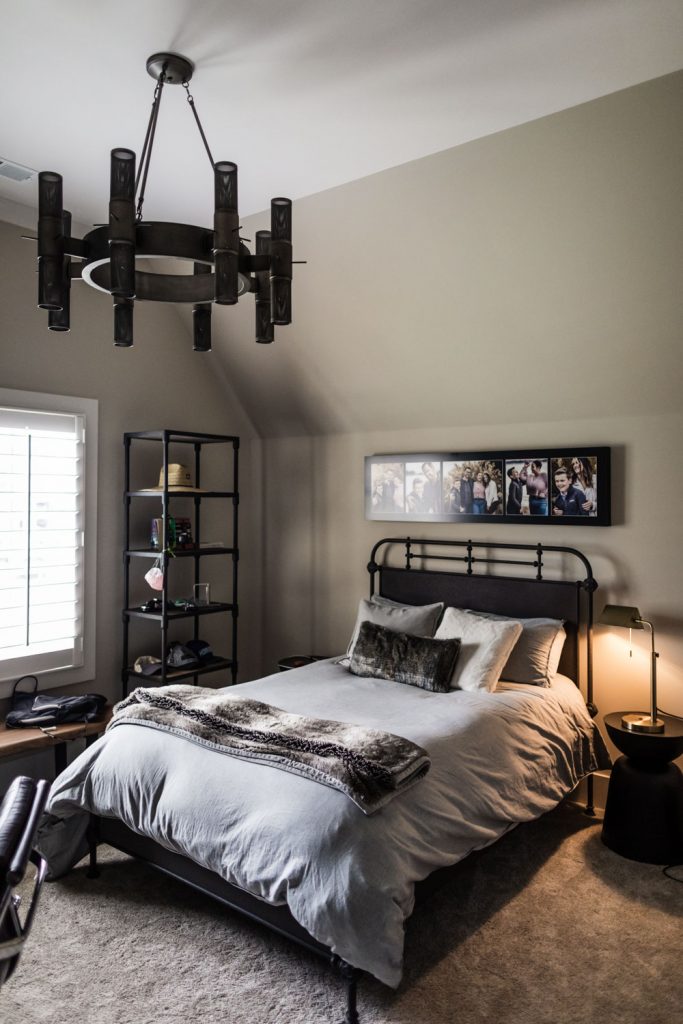A modern bedroom with a metal-frame bed, gray bedding, pillows, and a fur throw. A tall black shelf stands in the corner, and a black chandelier hangs from the ceiling. Family photos are framed above the bed.
