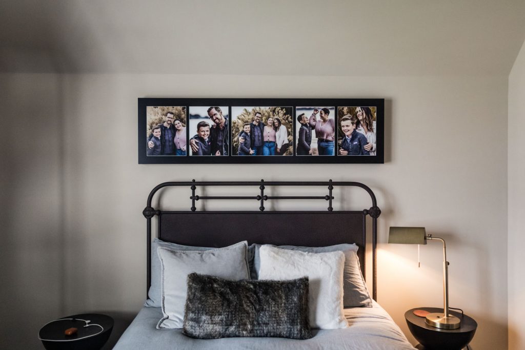 A neatly made bed with gray and white bedding sits beneath a series of five framed photos above the headboard. There are round side tables with a lamp and items on either side of the bed against a pale wall.
