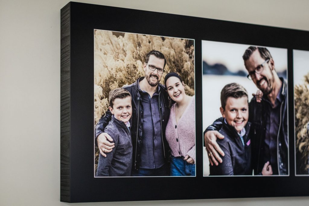 A framed photo collage on a wall shows a smiling man, woman, and boy standing closely together outdoors, with dried tall grasses in the background. Another photo features similar subjects in a different pose.