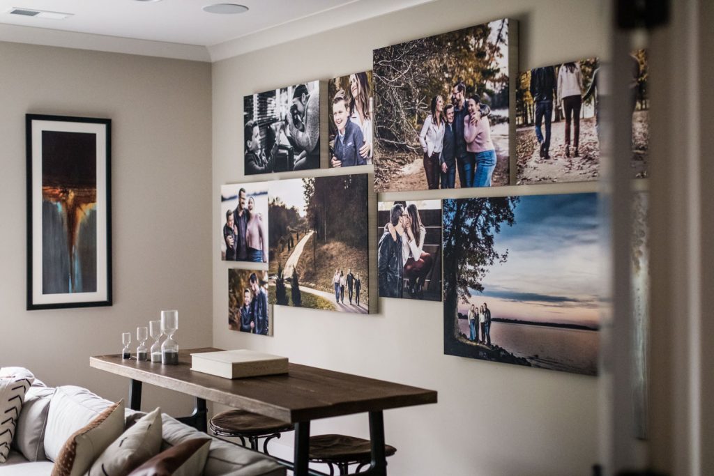 A living room with a wooden table, candles, and a couch. The wall displays several large, framed family photos in various outdoor settings, along with a single abstract painting to the left.