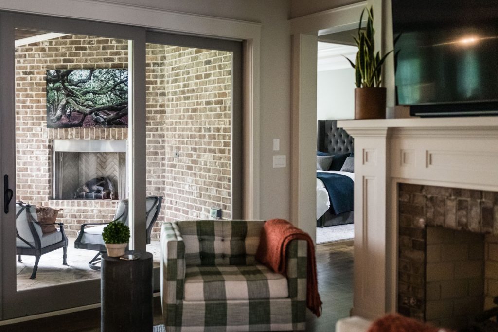 A cozy living room with a green plaid armchair, a small potted plant on a side table, and a white fireplace. Sliding glass doors open to a brick patio with chairs and a fireplace. A bedroom is visible in the background.