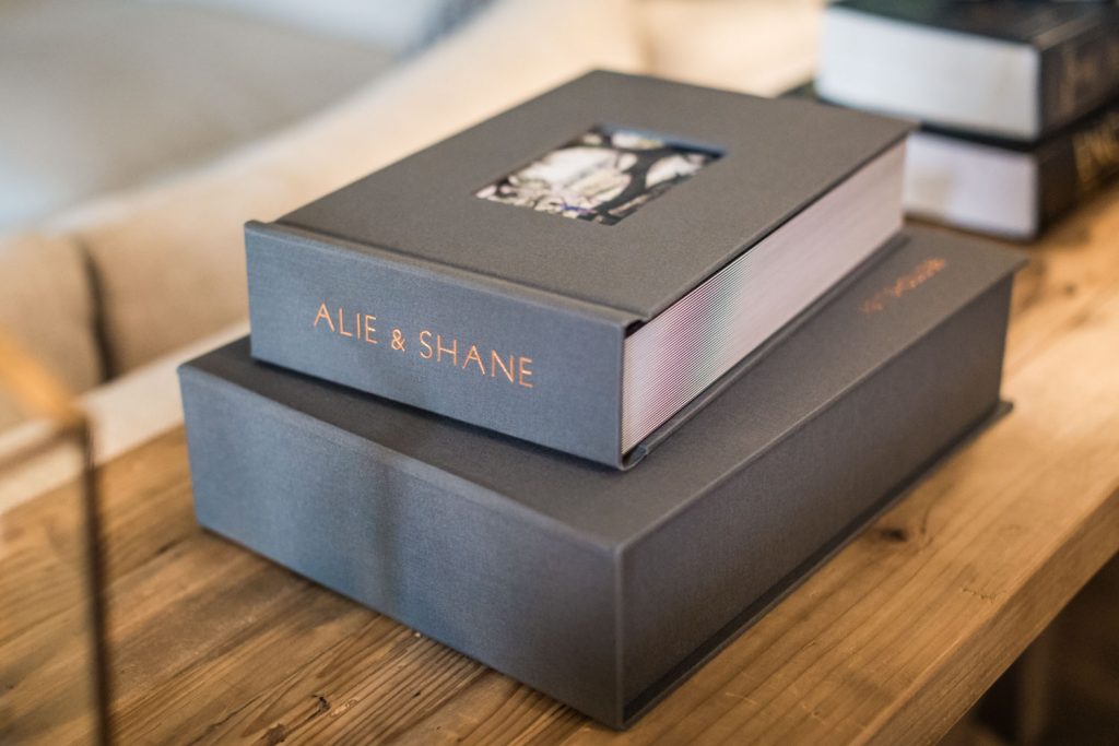 Two gray photo albums, one atop the other, with ALIE & SHANE embossed in gold on the spine, sit on a wooden table. A small photo is visible on the top albums cover. Other books are blurred in the background.