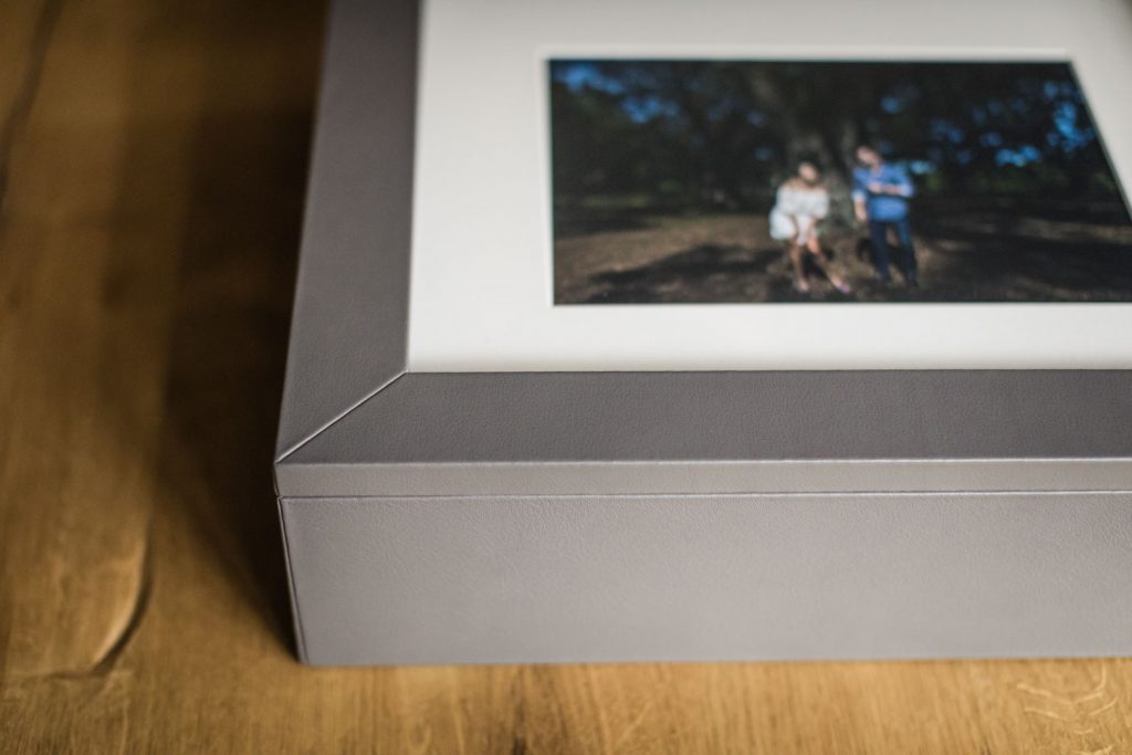 A close-up of a gray photo box with a framed photo on top, sitting on a wooden surface. The photo in the frame is slightly blurred and shows two people outdoors.
